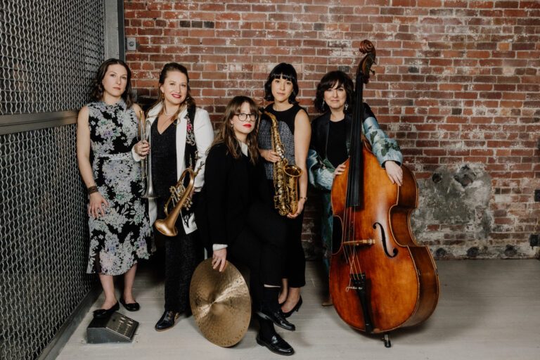A group of five women with instruments pose in front of a brick wall. 