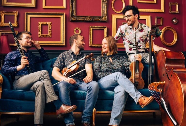 The members of Vinta sit on a cough, smiling at each other while displaying their instruments. 