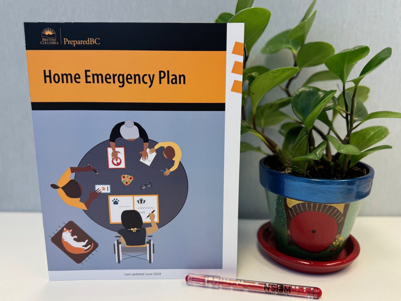 A copy of Home Emergency Plan sits on a desk beside a potted plant and a pen.