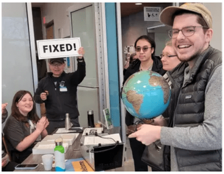 A group of volunteers stand around a table of broken items. One holds a globe and another holds up a sign that proclaims FIXED!